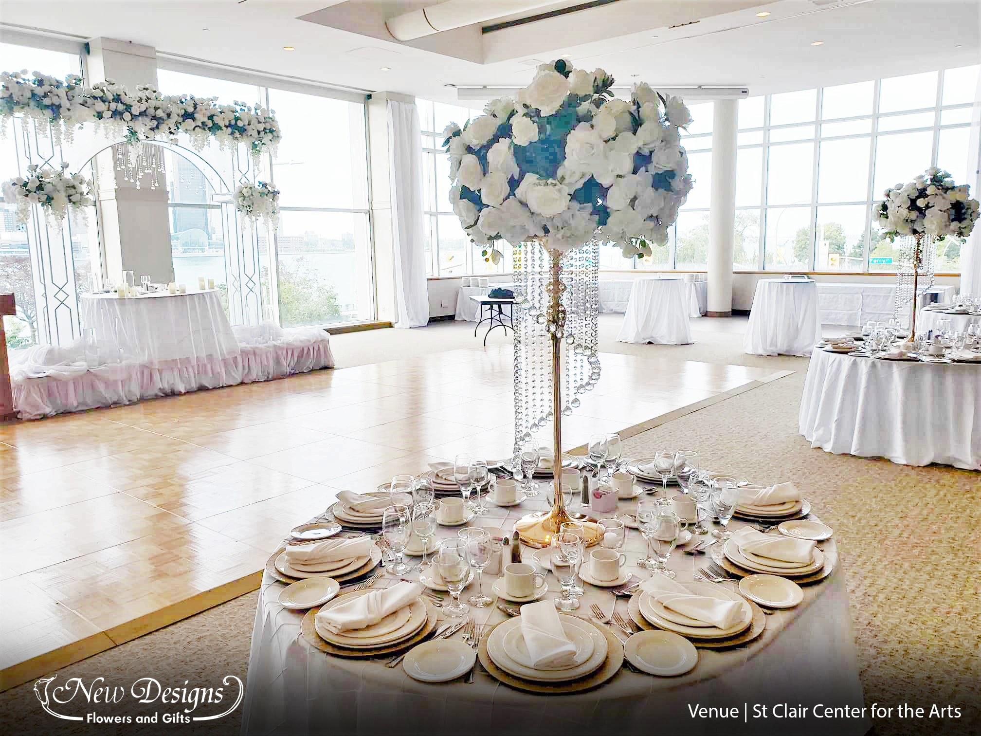 A hall overflowing with draped White Chiffon along the walls and on the tables, Elegantly pulled back to reveal an entry door. With a Tall 4 posted gold stand adorned on top with a full compact arrangement of white roses hydrangeas and orchids Against a full wall of windows facing the waterfront, sits a stunning 8ft tall white metal arch with frosted panels, overflowing with white roses and orchids